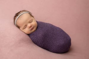 Newborn baby on bean nag wrapped in a purple wrap. 